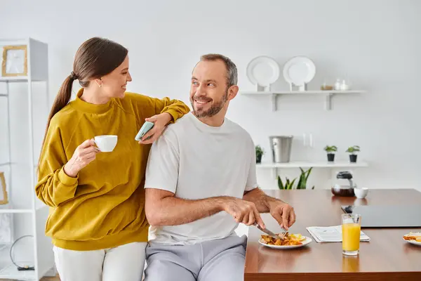 Cheerful man enjoying tasty breakfast near wife with coffee cup and smartphone in modern kitchen — Stock Photo