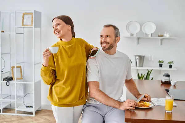 Hombre feliz disfrutando de sabroso desayuno cerca de la esposa con taza de café y teléfono inteligente mirando hacia otro lado en la cocina - foto de stock