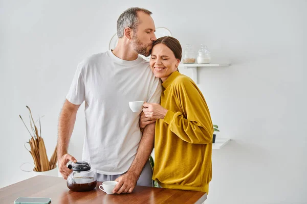 Husband kissing head of cheerful wife near fresh aromatic coffee in kitchen, child-free couple — Stock Photo