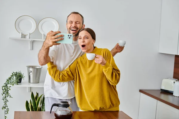 Счастливая пара без детей веселится и делает селфи во время утреннего кофе на современной кухне — стоковое фото