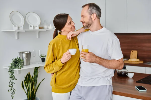 Feliz pareja sin niños con café y jugo de naranja fresco mirándose en la cocina - foto de stock