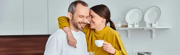Mujer cariñosa con taza de café de la mañana abrazando marido sonriente en la cocina, pancarta horizontal - foto de stock