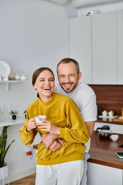 Cheerful man embracing laughing wife holding fresh morning coffee in kitchen, child-free concept — Stock Photo