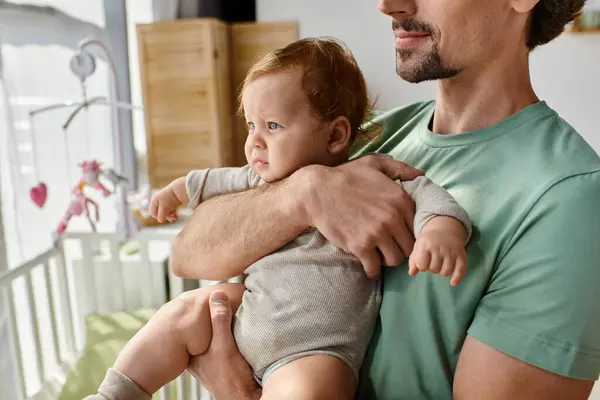 Tiro recortado de padre barbudo sosteniendo en brazos a su hijo pequeño en el acogedor dormitorio en casa, la paternidad - foto de stock