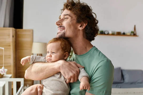 Dai capelli ricci e padre allegro che tiene in braccio il suo bambino in camera da letto accogliente a casa, paternità — Foto stock