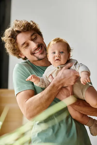 Cheveux bouclés et heureux père tenant dans les bras son fils bébé dans une chambre confortable à la maison, la paternité — Photo de stock