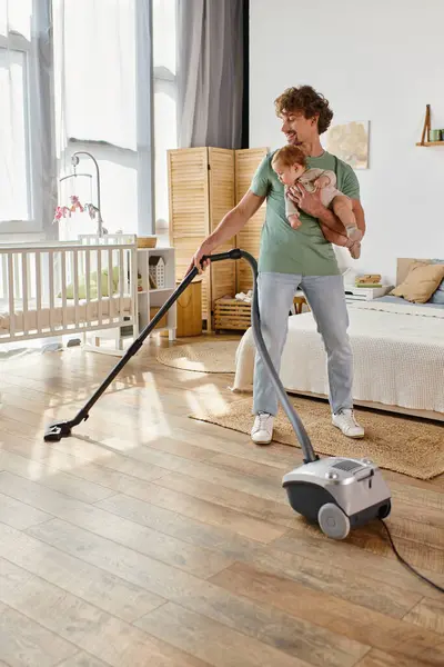 Frisé père multitâche ménage et garde d'enfants, homme aspirant chambre avec bébé fils dans les bras — Photo de stock