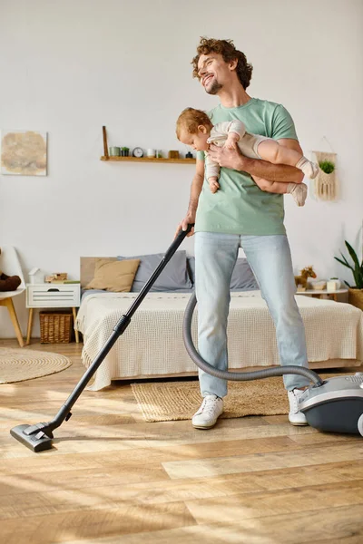 Homem multitarefa trabalho doméstico e puericultura, pai feliz aspirando apartamento com menino nos braços — Fotografia de Stock