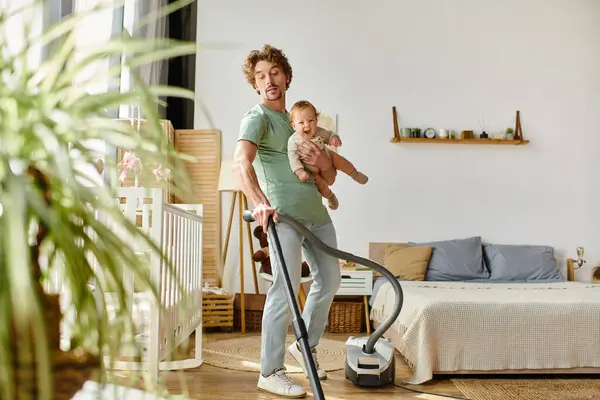 Homme multitâche travaux ménagers et de garde d'enfants, bouclé père aspirateur appartement avec bébé garçon dans les bras — Photo de stock