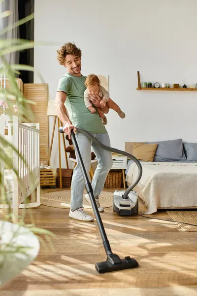 Hombre multitarea tareas tareas domésticas y cuidado de niños, sonriente padre aspiradora apartamento con niño de brazos - foto de stock