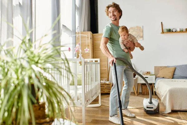 Hombre multitarea tareas tareas domésticas y cuidado de niños, apartamento de aspiradora de padre soltero con niño de brazos - foto de stock
