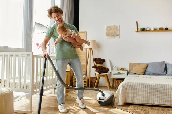 Homme multitâche travaux ménagers et de garde d'enfants, père aspirant plancher de bois franc avec bébé garçon dans les bras — Photo de stock