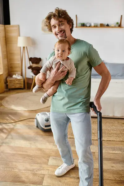 Happy man multitasking housework and childcare, father vacuum uming hardwood floor with son in arms — стоковое фото