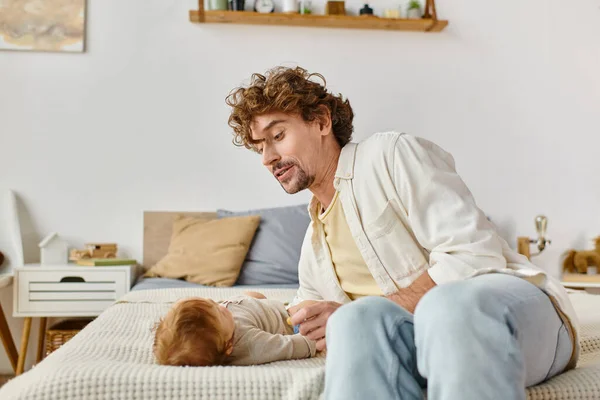 Père célibataire aux cheveux bouclés tenant la sucette près du bébé garçon sur le lit, la paternité et l'amour — Photo de stock