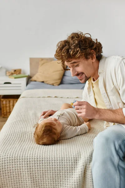 Père bouclé et barbu tenant la sucette près du bébé garçon sur le lit, la paternité et l'amour — Photo de stock