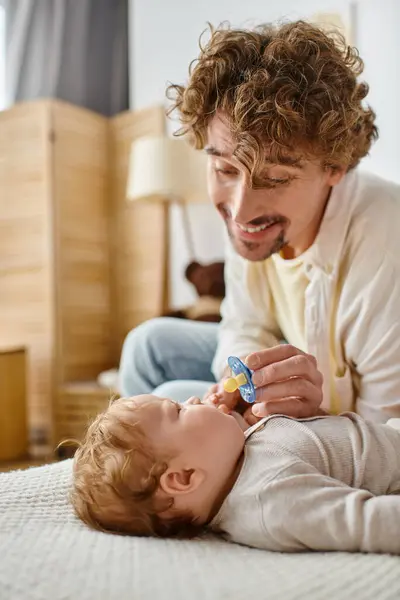 Heureux et frisé père donnant sucette à son fils en bas âge sur lit confortable, la paternité et l'amour — Photo de stock