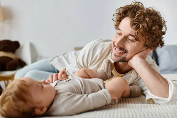 Pai alegre com cabelo encaracolado e barba olhando para seu filho enquanto deitado juntos na cama, amor — Fotografia de Stock
