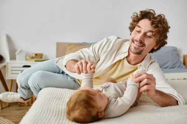 Happy father with curly hair holding hands of his baby boy while lying together on bed, innocent — Stock Photo