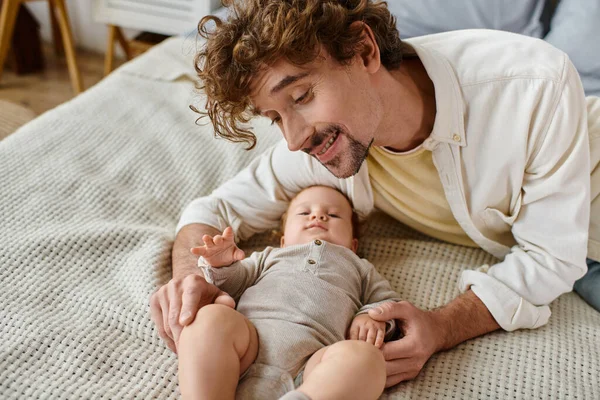 Alegre hombre con el pelo rizado y la barba mirando lindo bebé hijo en la cama, momentos preciosos - foto de stock