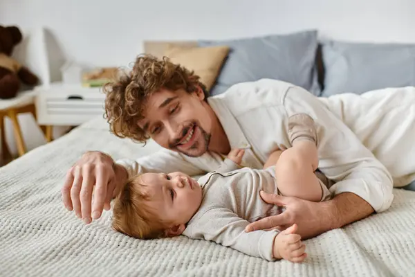 Pai alegre com cabelo encaracolado e barba olhando para seu filho bebê bebê na cama, momentos preciosos — Fotografia de Stock