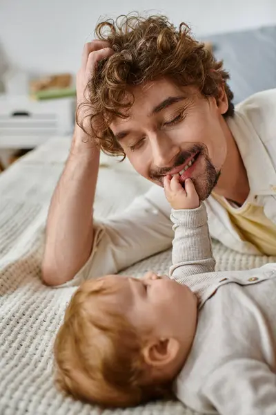 Lindo bebé niño acostado en la cama y tocando la cara de su padre feliz barbudo, momentos preciosos - foto de stock