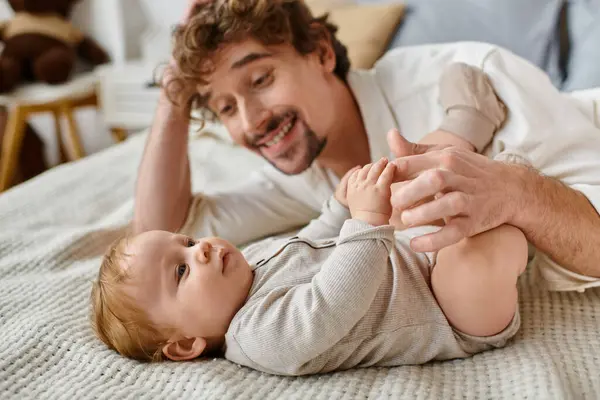 Cute infant baby boy lying on bed and holding hand of his bearded happy father, precious moments — Stock Photo