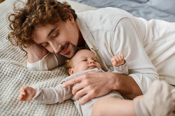 Homme heureux avec les cheveux bouclés embrassant son bébé tout en étant couché ensemble sur le lit, nourrissant et l'amour — Photo de stock