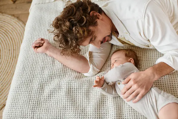Happy man with curly hair embracing his infant baby while lying together on bed, horizontal banner — Stock Photo