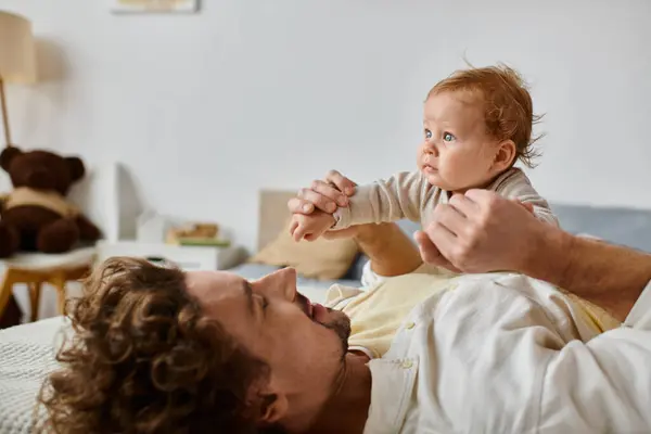Mann mit lockigem Haar und Bart hält Händchen mit seinem kleinen Sohn mit blauen Augen im Schlafzimmer — Stockfoto