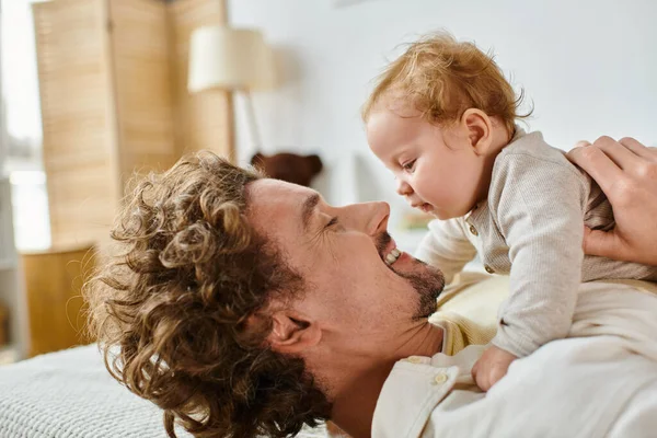 Homme heureux avec les cheveux bouclés et la barbe en regardant son fils en bas âge dans la chambre à coucher, l'amour et les soins du père — Photo de stock