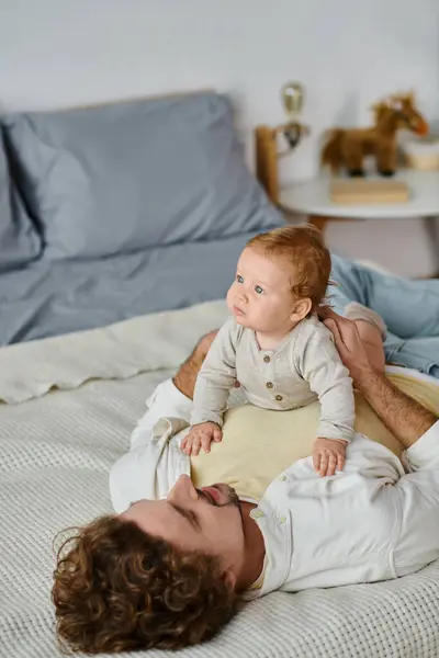 Homem com cabelo encaracolado e barba abraçando seu filho bebê em uma cama, ligação entre pai e filho — Fotografia de Stock
