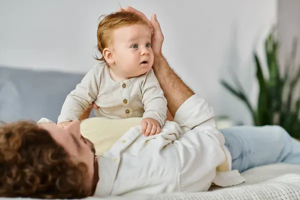 Curly man lying on bed with his cute infant son with blue eyes, bonding between father and child — Stock Photo