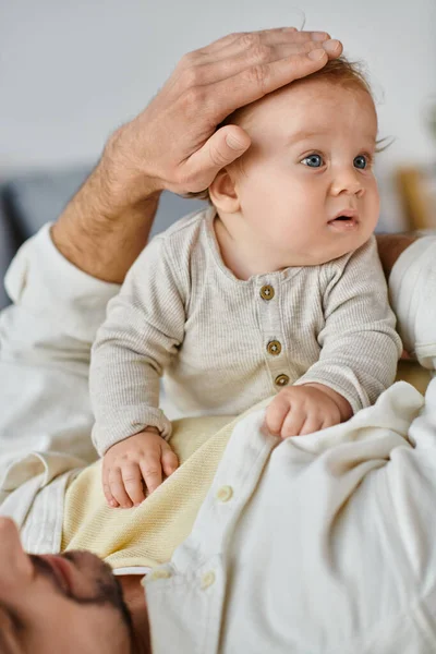 Alleinerziehender Vater mit lockigem Haar und Bart streichelt Haare seines kleinen Jungen im Schlafzimmer, Liebe und Fürsorge — Stockfoto