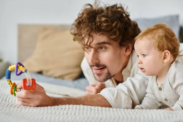 Père célibataire et bébé garçon captivé par hochet coloré dans la chambre à coucher, lien entre parent et enfant — Photo de stock