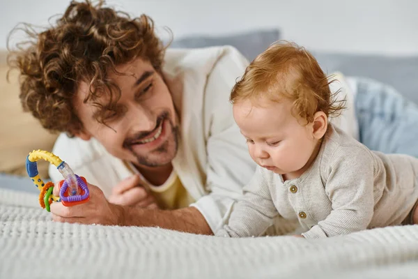 Père célibataire tenant hochet coloré près bébé garçon dans la chambre à coucher, lien entre père et fils — Photo de stock