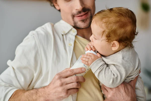 Обрізаний батько з бородою годує свого немовляти сином поживним молоком від дитячої пляшки, харчуючись — стокове фото