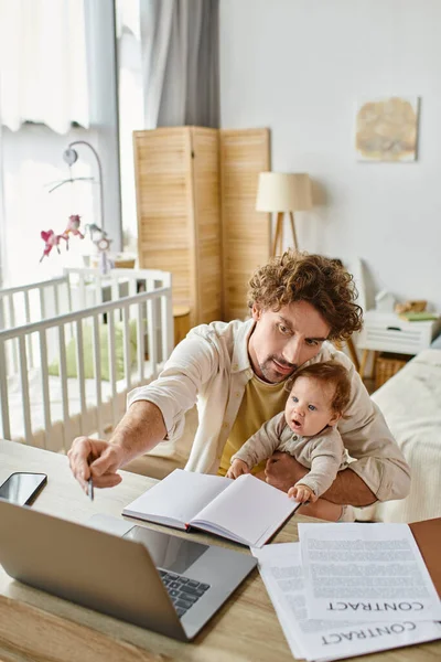 Батько тримає в руках свого немовляти сина, працюючи дистанційно на ноутбуці з дому, баланс робочого життя — стокове фото