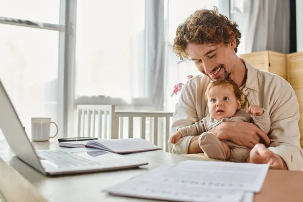 Счастливый отец держит младенца сына во время работы из дома рядом с бумагами и гаджетами, баланс работы и жизни — стоковое фото
