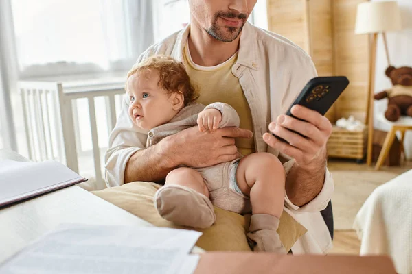 Uomo ritagliato che tiene il figlio durante l'utilizzo di smartphone e lavorare da casa vicino a carte e laptop — Foto stock