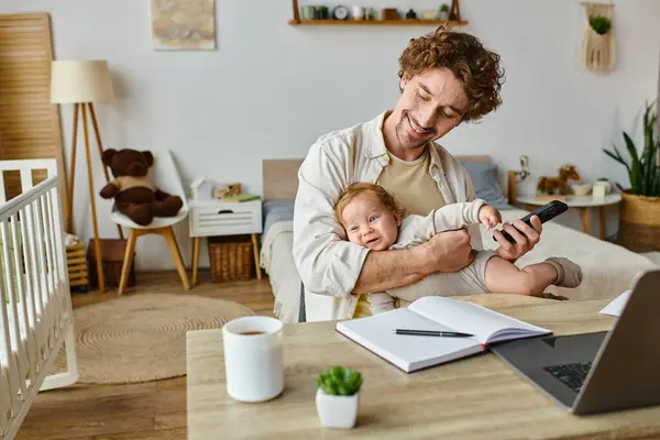 Uomo felice che tiene il bambino mentre tiene lo smartphone e lavora da casa vicino a documenti e laptop — Foto stock