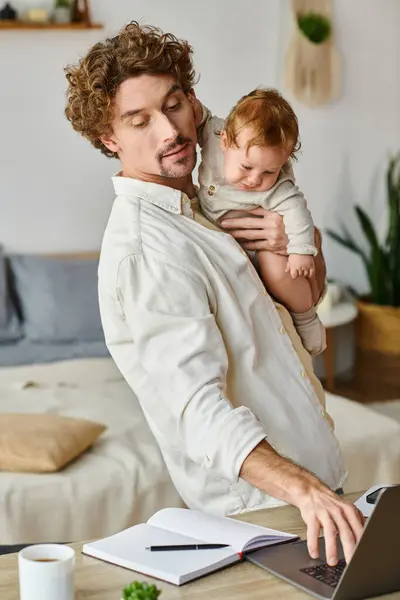 Щасливий і кучерявий батько тримає в руках свого немовляти сина і використовує ноутбук на столі, баланс робочого життя — стокове фото