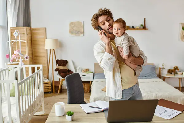 Мужчина держит младенца сына во время разговора на смартфоне и глядя на ноутбук на столе, работа из дома — стоковое фото