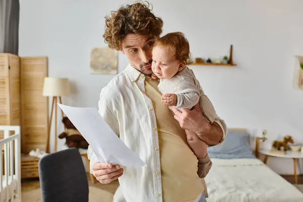 Padre riccio e barbuto che tiene in braccio il bambino neonato e guarda il documento, l'equilibrio della vita lavorativa — Foto stock