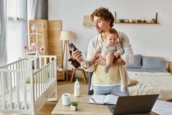 Занятой отец-одиночка, держащий младенца и использующий смартфон рядом с ноутбуком и детской бутылочкой на столе — стоковое фото