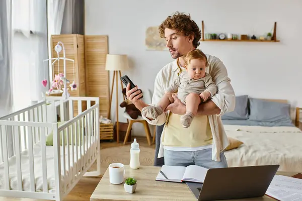 Padre single occupato che tiene in braccio il figlio neonato e utilizza smartphone vicino al computer portatile e al biberon sulla scrivania — Foto stock