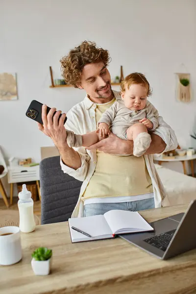 Щасливий батько тримає немовляти сина і використовує смартфон біля ноутбука і дитячої пляшки на столі — стокове фото