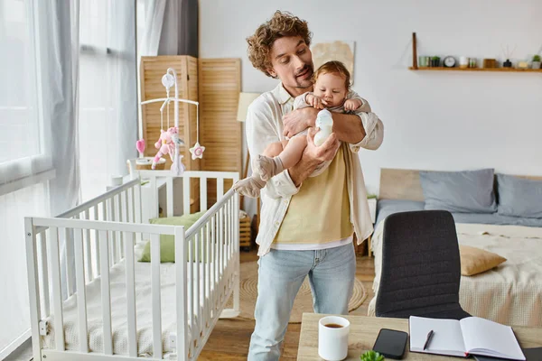 Cura padre single che alimenta il figlio neonato dal biberon mentre in piedi vicino alla culla e alla scrivania — Foto stock