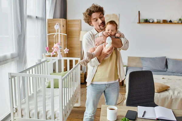 Caring single father feeding infant son from baby bottle while standing near crib and working desk — Stock Photo