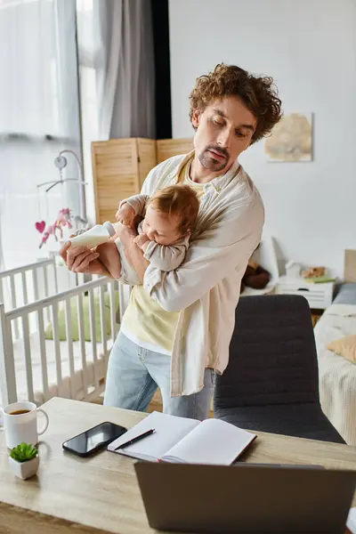 Один батько тримає сина і пляшку дитини, дивлячись на ноутбук на столі, баланс робочого життя — стокове фото