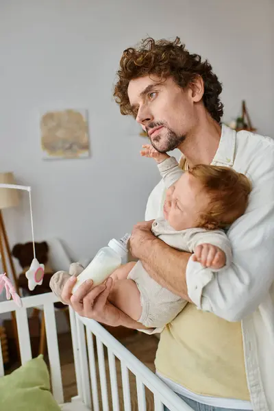 Curly-haired father feeding his infant son from baby bottle near crib, fatherhood and care — Stock Photo